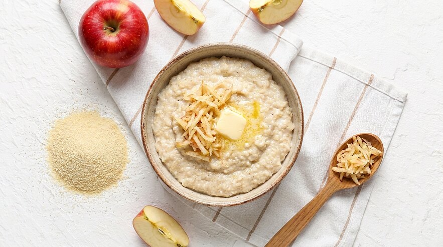 Schüssel mit warmem Grießbrei, garniert mit schmelzender Butter und geriebenem Apfel; daneben Apfelscheiben, ein ganzer Apfel und ein Löffel mit geriebenem Apfel.