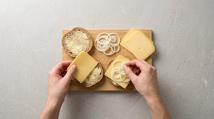 Von oben: Hände belegen aufgeschnittene Brötchen mit Käsescheiben; eine Hand legt Käse, die andere Zwiebelringe, alles auf einem Holzbrett.