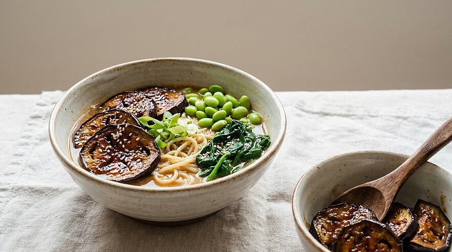 Eine Schüssel mit Ramen-Nudeln, garniert mit gebratenen Auberginenscheiben, grünen Edamame und frischem Spinat. Neben der Hauptschüssel steht eine kleinere Schüssel mit zusätzlichen gebratenen Auberginen. Der Hintergrund ist hell und schlicht gehalten.