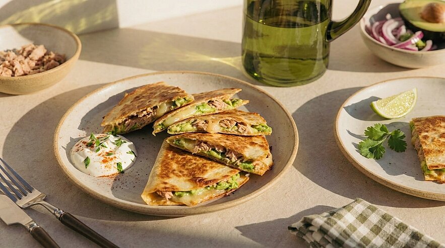 Goldbraune Quesadilla‑Dreiecke mit gezogenem Fleisch, Avocado und geschmolzenem Käse auf einem Teller, serviert mit Sauerrahm und Limettenspalte.