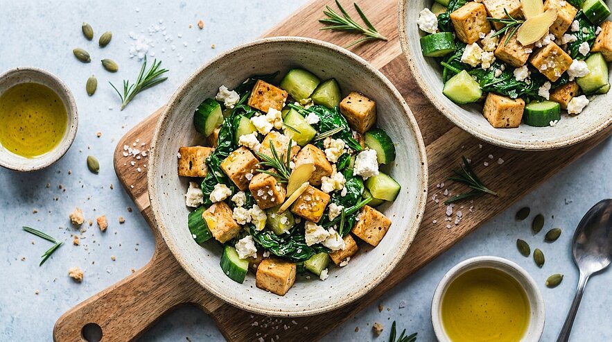 Zwei Schalen mit frischem Salat aus gebratenen Tofuwürfeln, Gurken, grünem Blattgemüse, zerbröseltem Feta und Rosmarin, dazu Olivenöl-Dressing.