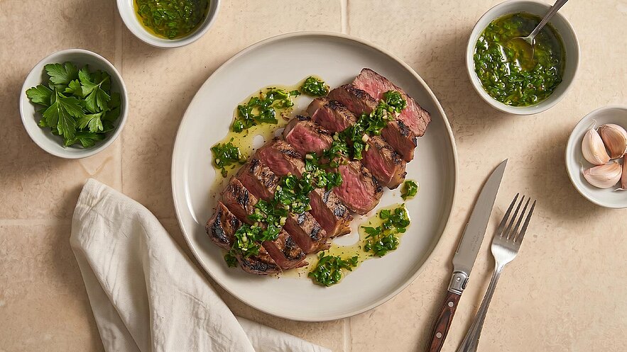 Auf dem Bild liegt ein aufgeschnittener Steak auf einem Teller, umgeben von grüner Sauce und frischen Kräutern. Neben dem Teller stehen mehrere kleine Schalen mit einer ähnlichen Sauce und Kräutern sowie einer Knoblauchknolle. Ein Besteckset liegt bereit.