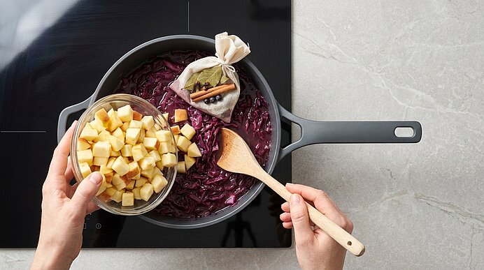 Hände geben gewürfelte Äpfel aus einer Glasschüssel in einen Topf mit Rotkohl auf dem Herd; ein Holzlöffel rührt, im Rotkohl liegt ein Gewürzbeutel mit Zimtstange.