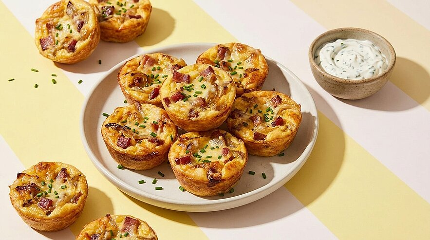 Teller mit mehreren herzhaften Mini-Quiches (Eimuffins) mit Speck und Schnittlauch, daneben eine kleine Schale mit Kräuter-Dip.