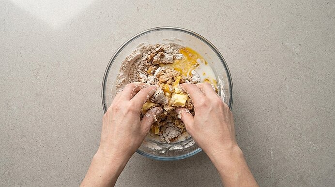 Zwei Hände kneten Teig in einer Glasschüssel; Mehl, aufgeschlagene Eier und Butter werden vermischt.