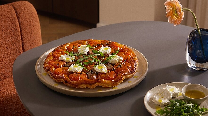 Herzhafte Tarte mit karamellisierten Tomaten, garniert mit kleinen Häufchen Frischkäse und Rucola, auf einem Teller serviert.
