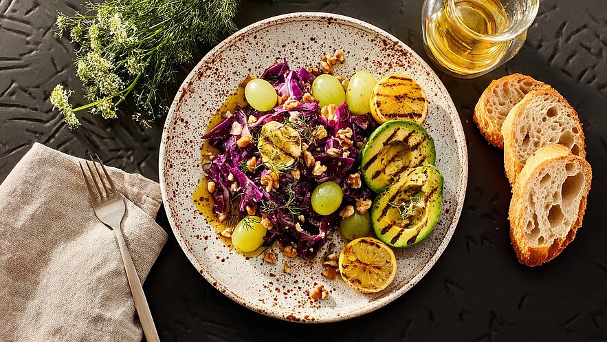 Eine Runde Schale mit lila Krautsalat, grünen Trauben und gehackten Walnüssen. Gegrillte Avocado und Zitrone ergänzen das Gericht. Daneben liegen einige Brotscheiben und ein Glas Öl. Der Hintergrund ist dunkel.