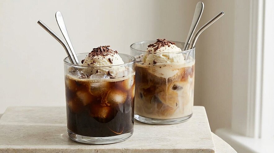 Zwei Gläser Eiskaffee mit Eiswürfeln, jeweils eine Kugel Vanilleeis und Schokoraspel obenauf; ein Getränk dunkel, das andere mit Milch aufgegossen. In jedem Glas steckt ein langer Löffel.