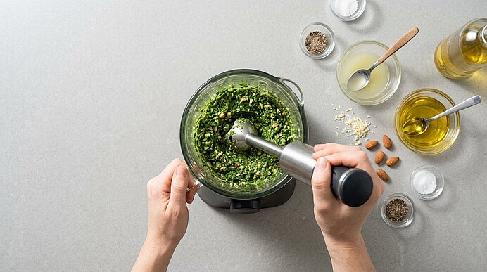 Zwei Hände pürieren Rucola, Parmesan und Mandeln in einem Glasgefäß mit einem Stabmixer zu grünem Pesto.