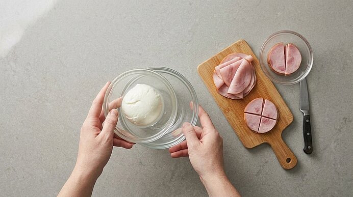 Zwei Hände halten eine Schüssel mit Mozzarella. Daneben liegen geschnittene Schinkenscheiben auf einem Holzbrett.