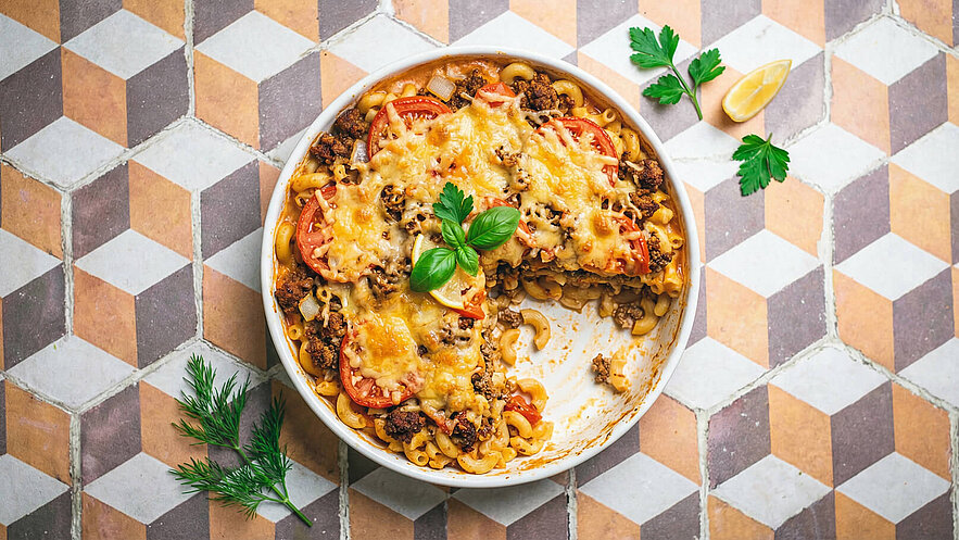 Ein Auflauf aus Makkaroni, Hackfleisch und Tomaten ist dekorativ in einer runden Auflaufform angerichtet. Der Auflauf ist goldbraun überbacken und mit frischen Kräutern, darunter ein Basilikumblatt, garniert. Teile des Auflaufs wurden bereits entnommen.