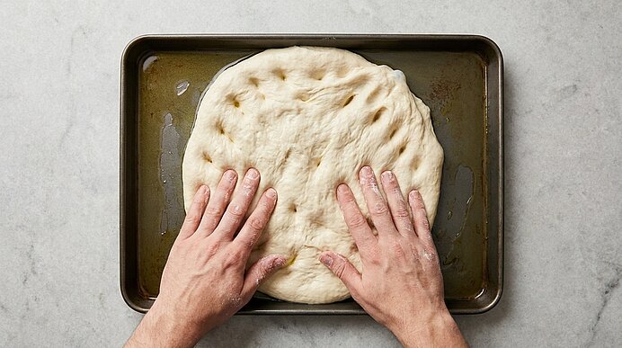 Zwei Hände drücken den Pizzateig mit den Fingerspitzen auf einem geölten dunklen Backblech flach.