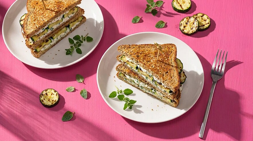 Zwei getoastete Sandwichs, jeweils halbiert, gefüllt mit gegrillten Zucchinischeiben, Frischkäse und frischen Kräutern, auf weißen Tellern auf pinkem Untergrund, neben einer Gabel.