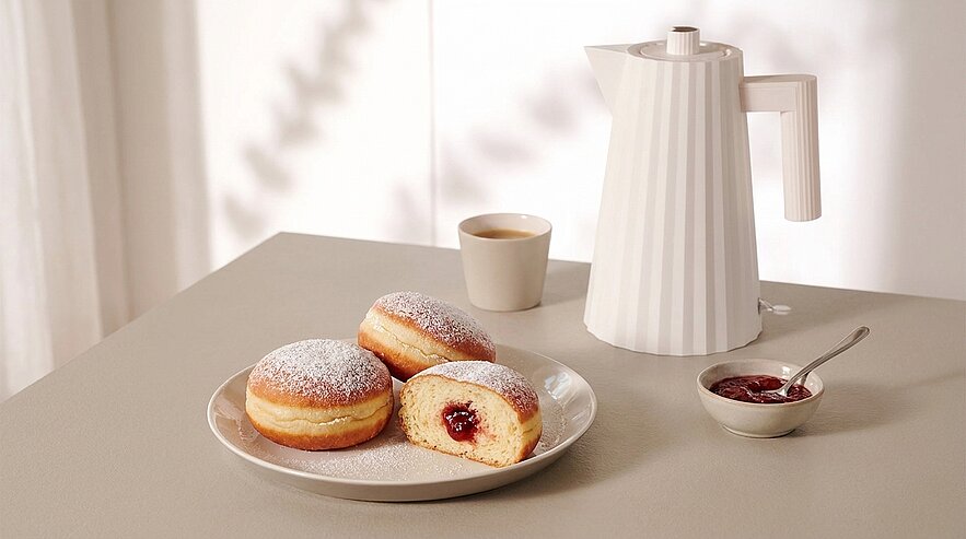 Drei mit roter Konfitüre gefüllte Krapfen auf einem Teller, einer halbiert und die Füllung sichtbar, bestäubt mit Puderzucker; daneben Tasse Kaffee, Thermoskanne und kleine Schale mit Marmelade und Löffel.