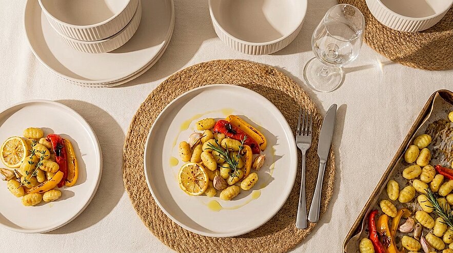 Teller mit gerösteten kleinen Kartoffeln, Zitronenscheiben, bunten Paprikastreifen und Rosmarinzweigen, mit Olivenöl beträufelt; daneben Besteck und ein Blech mit weiteren Kartoffeln.