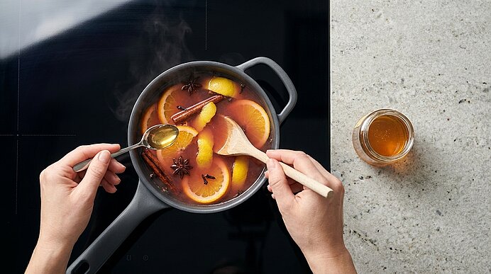 Zwei Hände rühren in einem Topf heißen Gewürz-Punsch mit Orangenscheiben, Zimtstangen und Sternanis; ein Glas Honig steht daneben.