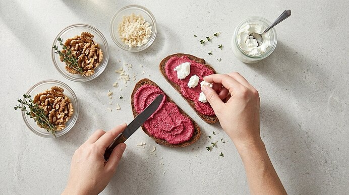 Zwei Hände belegen Brotscheiben mit rosa Rote-Rüben-Aufstrich und Ziegenfrischkäse auf einer hellgrauen Arbeitsplatte.