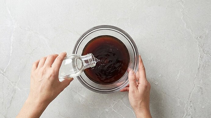 Vogelperspektive auf zwei Hände, die aus einer kleinen Glasflasche klare Flüssigkeit in eine Glasschüssel mit dunkler Flüssigkeit gießen.
