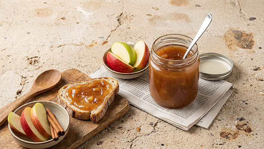 Ein rustikales Holzbrett zeigt einen frisch bestrichenen Toast mit Apfelmarmelade. Daneben liegt eine Schale mit Apfelscheiben und Zimtstangen. Im Hintergrund steht ein Glas mit der Marmelade und einem Löffel. Die Szene vermittelt ein gemütliches Frühstück.