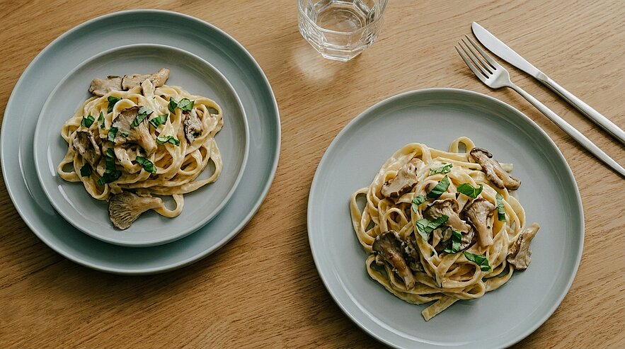 Zwei Teller mit Pasta auf einem Holztisch. Die Nudeln sind mit einer cremigen Sauce und Pilzen garniert. Frische Kräuter sind darüber verteilt. Neben den Tellern steht ein Glas Wasser und Besteck ist gut sichtbar.