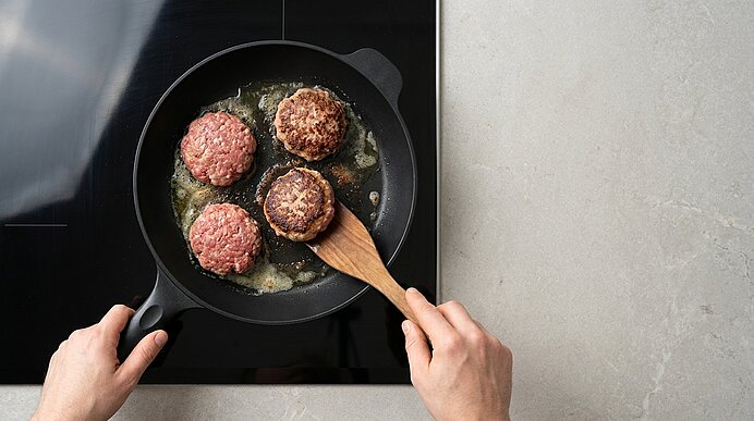 Zwei Hände braten Frikadellen in einer schwarzen Pfanne auf dem Kochfeld und wenden eine davon mit einem Pfannenwender.