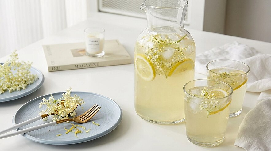 Eine Glas Karaffe mit einem erfrischenden Getränk, das mit Zitronen und Holunderblüten dekoriert ist, steht auf einem Tisch. Daneben befinden sich zwei Gläser mit dem gleichen Getränk sowie ein Teller mit einer Gabel und weiteren Holunderblüten.