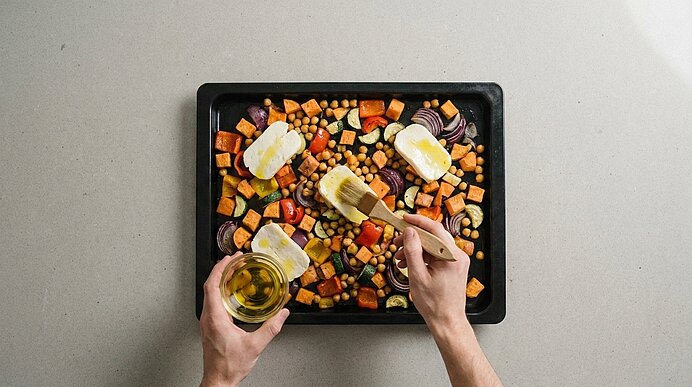Backblech mit bunt gewürfeltem Ofengemüse und Kichererbsen, vier Scheiben weichen Käses darauf; Hände pinseln Olivenöl über den Käse.