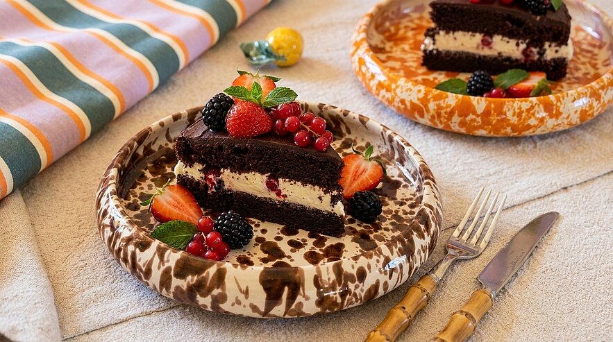 Auf einem Tisch liegen zwei Teller mit Stücke von Schokoladentorte. Die Torte ist mit Sahne, frischen Beeren und Minzblättern dekoriert. Im Hintergrund sieht man ein gestreiftes Tuch. Die Atmosphäre ist einladend und festlich.