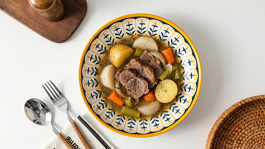 Ein Teller mit Hackfleisch und Gemüse, darunter Kartoffeln, Karotten und Sellerie, ist zentral abgebildet. Der Teller hat ein dekoratives Muster. Neben dem Teller liegen Besteck und eine Serviette. Im Hintergrund befindet sich ein Holzobjekt.