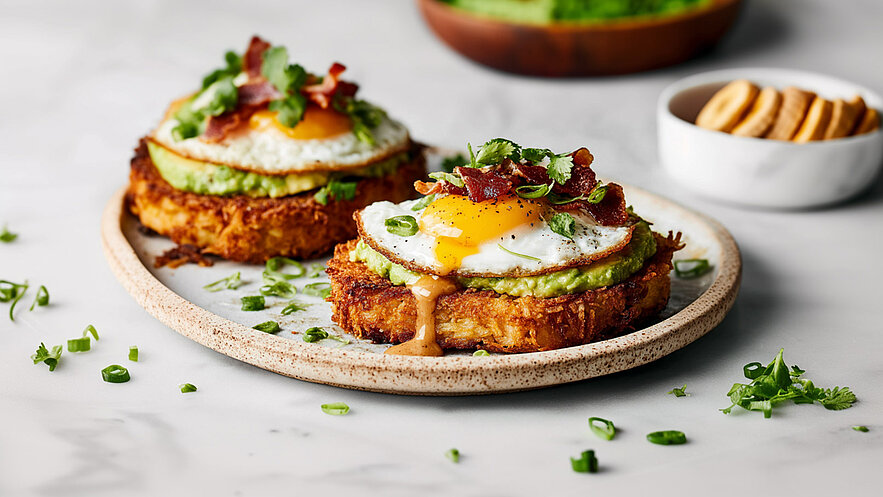 Ein Teller mit zwei goldbraun gebratenen Kartoffelröstis, belegt mit Avocado, Spiegelei und knusprigem Speck. Frischer Koriander und Frühlingszwiebeln garnieren das Gericht. Kleine grüne Kräuterstücke sind um die Röstis verteilt. Im Hintergrund ist eine Schüssel mit Keksen sichtbar.