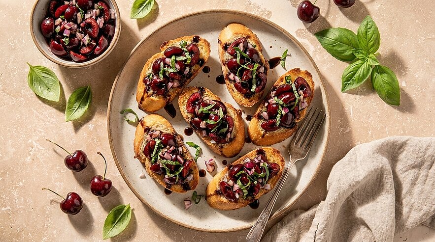 Sechs geröstete Crostini auf einem Teller, belegt mit gehackten Kirschen, frischem Basilikum und Balsamico-Glasur.
