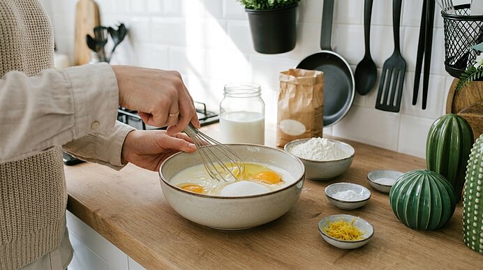 Hand einer Person rührt mit Schneebesen Eier in einer Schüssel auf einer Küchenarbeitsplatte, daneben Schalen mit Mehl, Zucker, Milch und Zitronenschale; Kochutensilien hängen an der Wand.