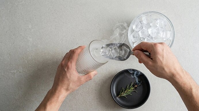 Zwei Hände füllen mit einer Eiskelle ein Glas mit Eiswürfeln.