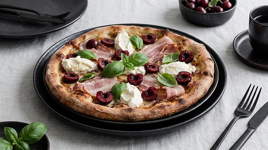 Nahaufnahme einer rustikalen Pizza mit knusprigem Rand, belegt mit Schinken, halbierten Kirschen, cremigem Burrata-Mozzarella und frischen Basilikumblättern auf einem schwarzen Teller.