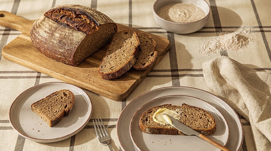 Rustikales Vollkornbrot auf einem Holzbrett mit zwei Scheiben; auf einem Teller eine Scheibe mit Butter bestrichen und Messer, daneben ein kleiner Teller mit weiterer Scheibe.