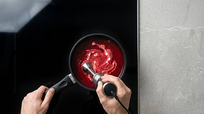 Zwei Hände pürieren Rote-Bete-Suppe mit Sahne mit einem Pürierstab in einem schwarzen Stieltopf auf dem Kochfeld.