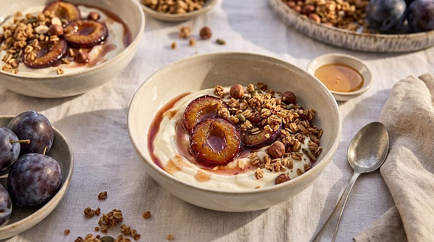 Schale mit Joghurt, karamellisierten Pflaumenhälften, knusprigem Granola und Sirup; weitere Pflaumen und ein Löffel daneben.