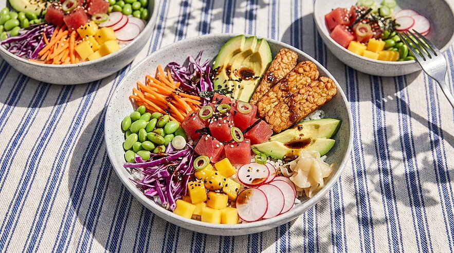 Drei bunte Poké-Bowls mit Würfeln rohem Thunfisch, Avocado, Tempeh, Edamame, Mango, Rotkohl, Karotten, Radieschen und Sesam, mit Soße serviert.