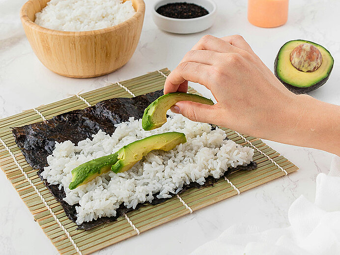 Eine Hand legt Avocadoscheiben auf eine Schicht aus Reis, die auf einem Nori-Blatt liegt. Im Hintergrund ist eine Schüssel mit Reis sichtbar. Das Bild zeigt die Zubereitung von Sushi oder ähnlichen Gerichten.