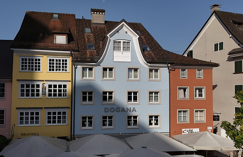 Farbenfrohe Gebäude in einer Stadtansicht, darunter ein blaues Haus mit dem Schriftzug "DOGANA". Die Architektur zeigt typische europäische Fassaden mit Fenstern und schrägen Dächern. Einige Terrassenschirme sind im Vordergrund sichtbar. Der Himmel ist klar und blau.
