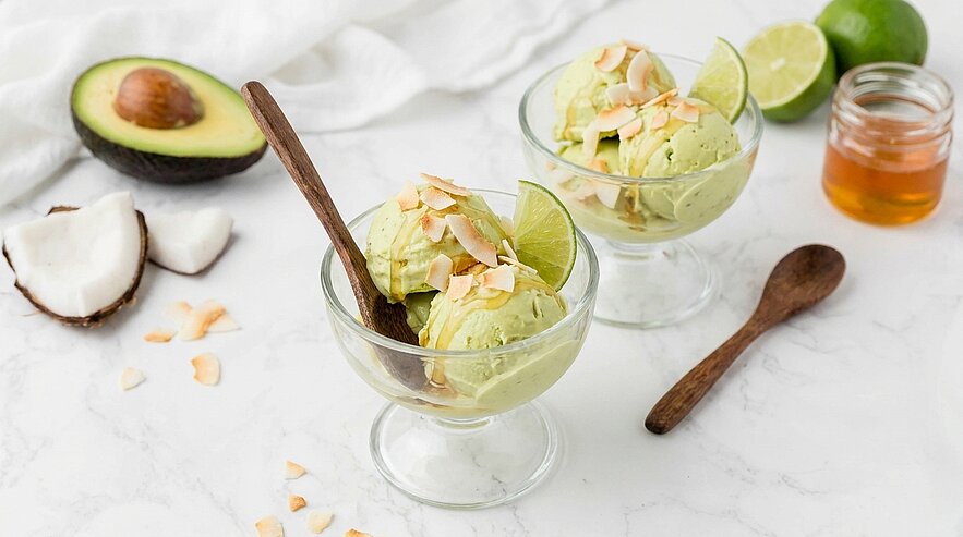 Zwei Glaschalen mit grüner Avocado-Limetten-Eiscreme, garniert mit Limettenscheiben und gerösteten Kokosflocken; daneben Holzlöffel und halbierte Avocado.