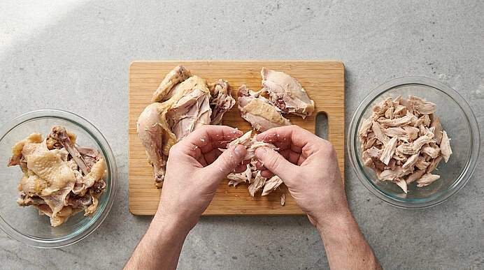 Zwei Hände zupfen gekochtes Hähnchenfleisch auf einem Holzbrett; links eine Schüssel mit Knochen, rechts eine Schüssel mit zerkleinertem Fleisch.