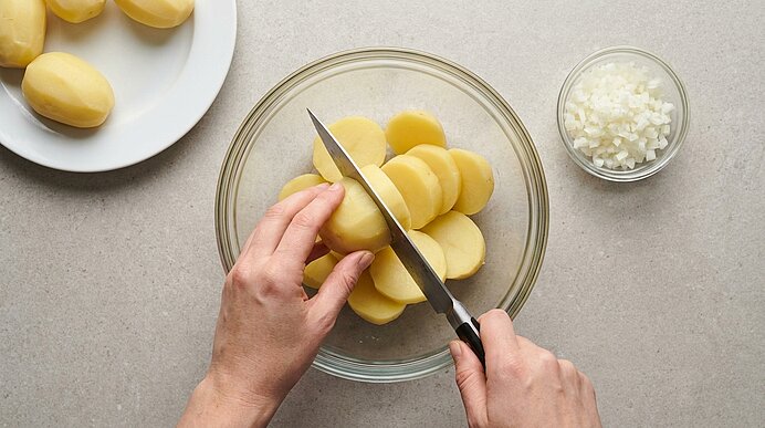 Zwei Hände schneiden geschälte Kartoffeln mit einem Messer in dünne Scheiben über einer Glasschüssel.