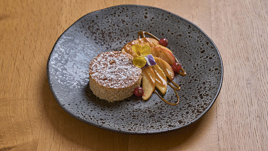 Ein fein dekorierter Dessertteller mit einem runden Kuchen, der mit Puderzucker bestäubt ist. Neben dem Kuchen sind orangefarbene Apfelscheiben, Beeren und eine süße Soße angerichtet. Eine essbare Blüte verleiht dem Gericht eine farbliche Akzentuierung.