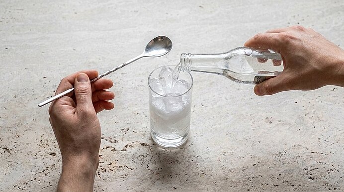 Rechte Hand gießt sprudelndes Wasser aus einer Glasflasche in ein Glas mit Eiswürfeln, linke Hand hält einen langen Löffel über dem Glas.