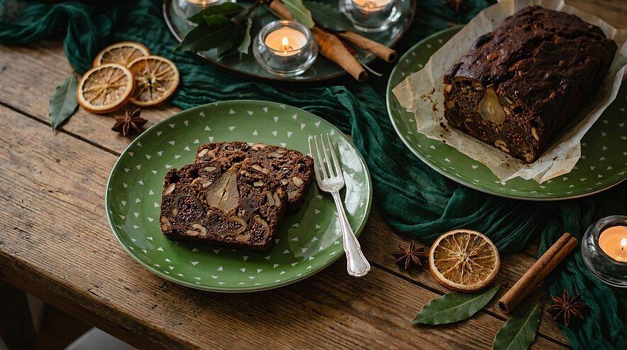 Scheibe dunkles Früchte-Nuss-Brot mit eingebackener Birne auf grünem Teller; daneben ganzes Brot, Zimtstangen und Kerzen auf rustikalem Holz­tisch.