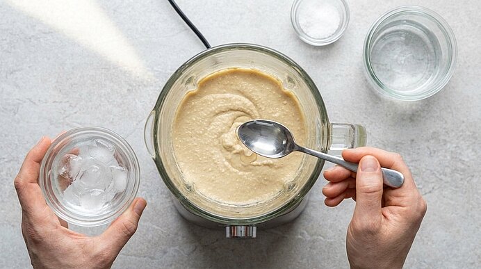 Zwei Hände geben mit einem Löffel Eiswasser aus einer Glasschale in einen Mixer mit hellem Hummus auf grauer Steinplatte.