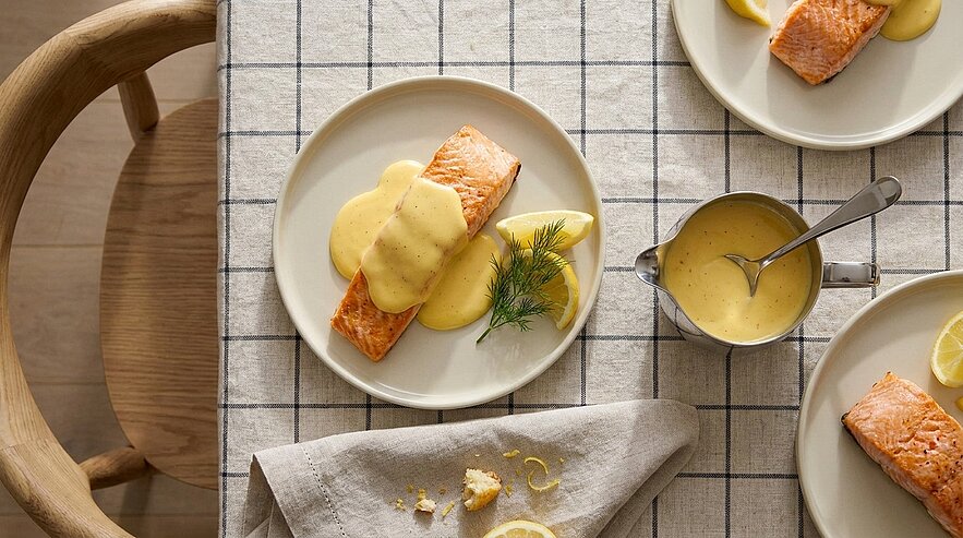 Gebratenes Lachsfilet auf einem Teller, übergossen mit cremiger Zitronen‑Hollandaise, garniert mit Dill und Zitronenscheibe; kleine Saucière daneben auf karierter Tischdecke.