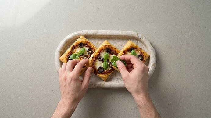 Zwei Hände garnieren gebackene Mini-Blätterteig-Pizzen auf einer Servierplatte mit frischen Basilikumblättern.