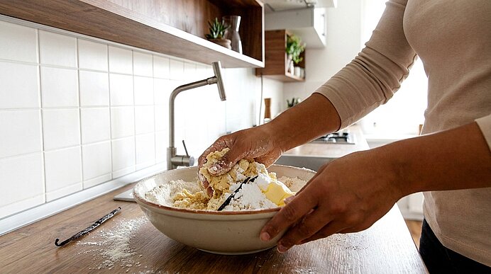 Hände einer Person, die in einer Schüssel Mehl und Butter mit einem Teigschaber zu Teig verarbeiten; Arbeitsfläche und Schüssel sind mit Mehl bestäubt.